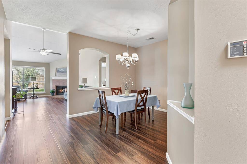 This photo showcases a bright, open-concept living and dining area. The space features elegant laminate wood floors, a modern chandelier above the dining table, and a cozy but spacious living room with a ceiling fan and fireplace. Large windows allow plenty of natural light. NOTE: Fireplace is virtually lit.