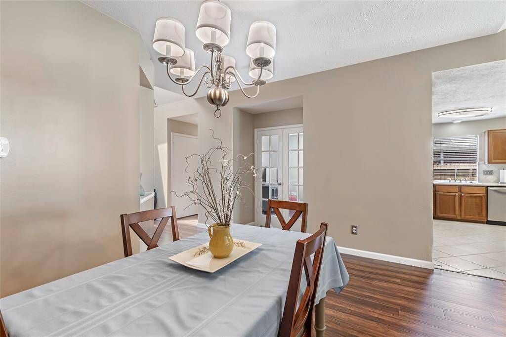 The dining area has a modern chandelier and features warm laminate wood flooring and neutral walls, leading to a bright kitchen with wooden cabinets and tile flooring, visible through a doorway.