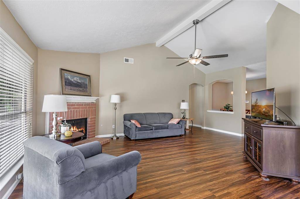 Another view of living room which features a cozy brick fireplace and vaulted ceiling with a fan. It has warm laminate wood flooring and neutral walls, creating an inviting atmosphere. NOTE: Fireplace is virtually lit.