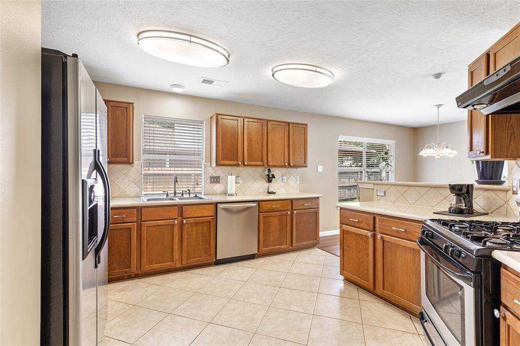 This kitchen features stainless steel appliances, wooden cabinets, and tiled flooring. It has ample Silestone counter space and Travertine backsplash with a window above the sink for natural light, and an adjacent dining area with a chandelier.