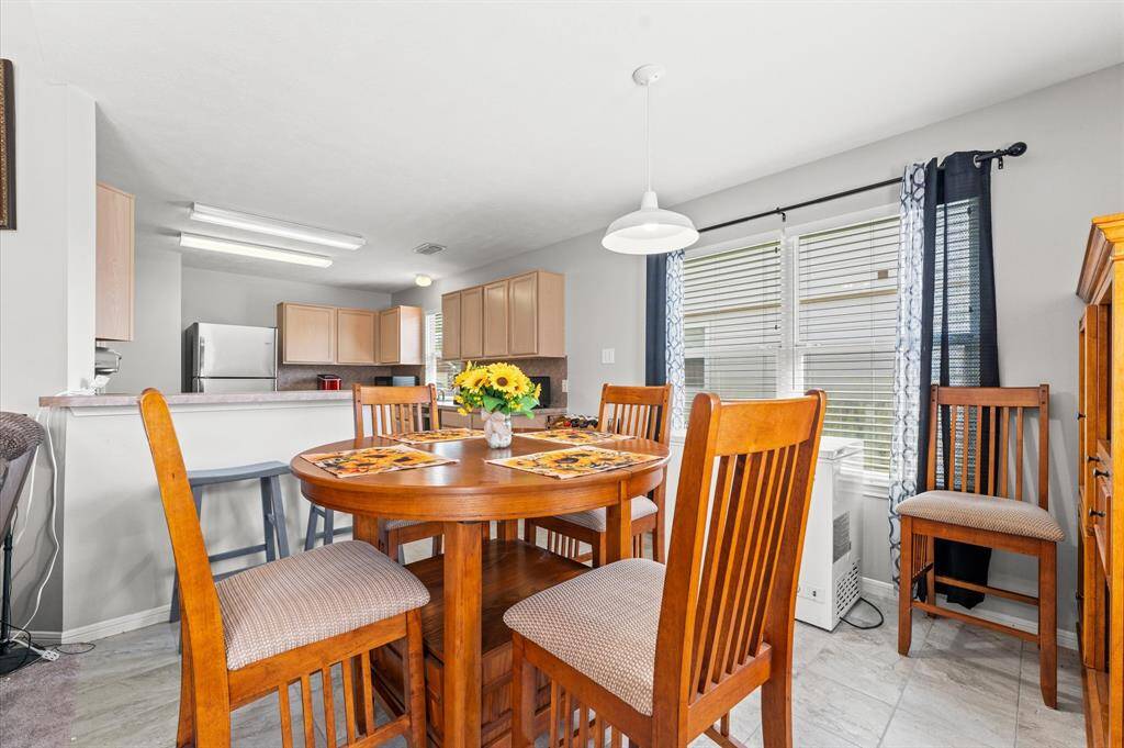 This photo shows a bright dining area adjacent to a kitchen with light cabinetry and stainless steel appliances. Large windows let in natural light, enhancing the cozy, welcoming atmosphere.