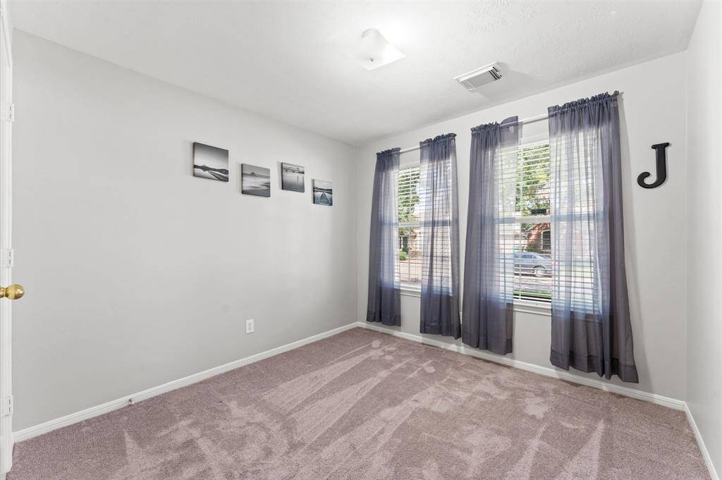This room features neutral walls, plush carpeting, and ample natural light from large windows with sheer curtains. Ideal for a bedroom or office space.