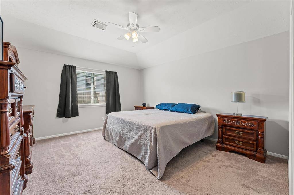 This bedroom features neutral walls and carpet, offering a cozy atmosphere. It includes a ceiling fan, a large window with dark curtains,  creating a classic and comfortable space.