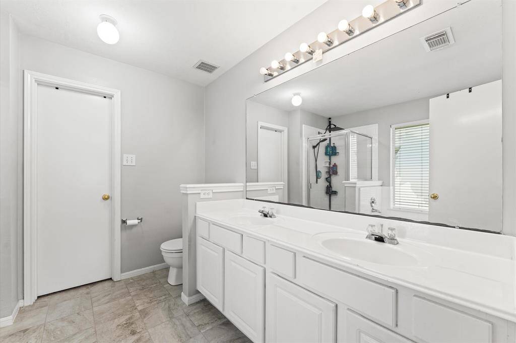 This bright bathroom features a large mirror, dual sinks with ample counter space, a walk-in shower, and a separate toilet area. The neutral tones and modern lighting create a clean, spacious feel.