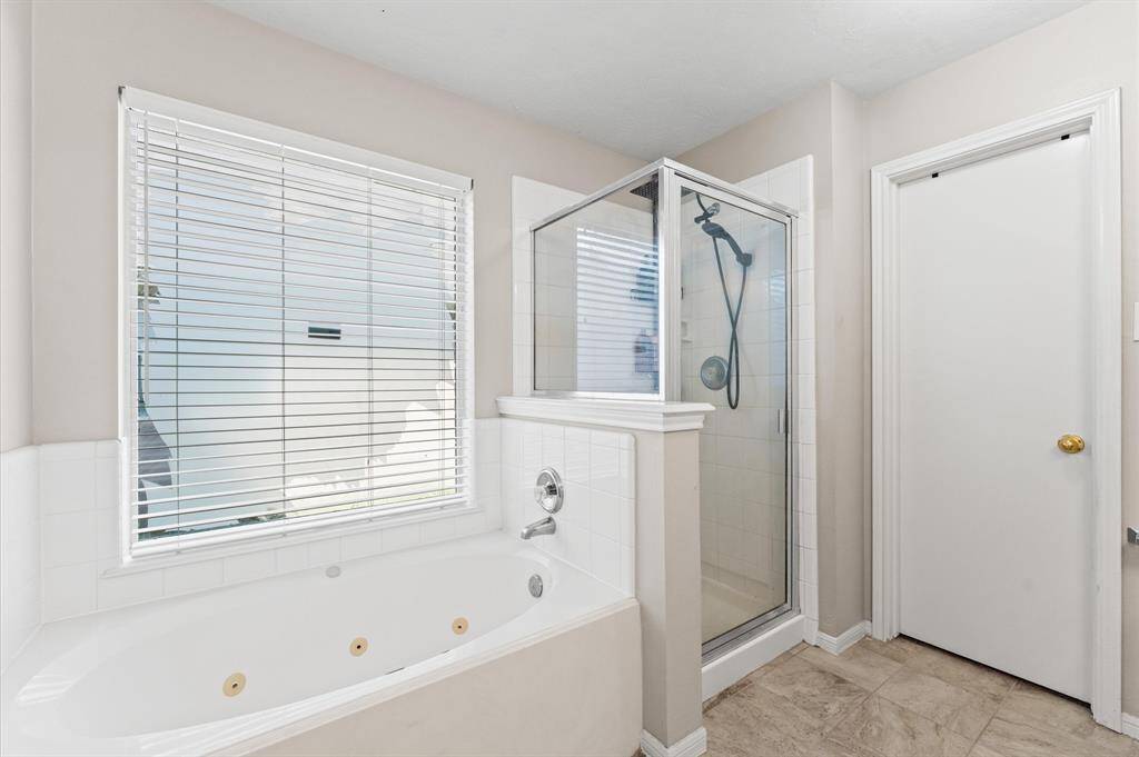 This bathroom features a spacious walk-in shower with glass enclosure, a large bathtub with jets, and a window providing natural light. The neutral tiles and walls offer a clean, modern feel.