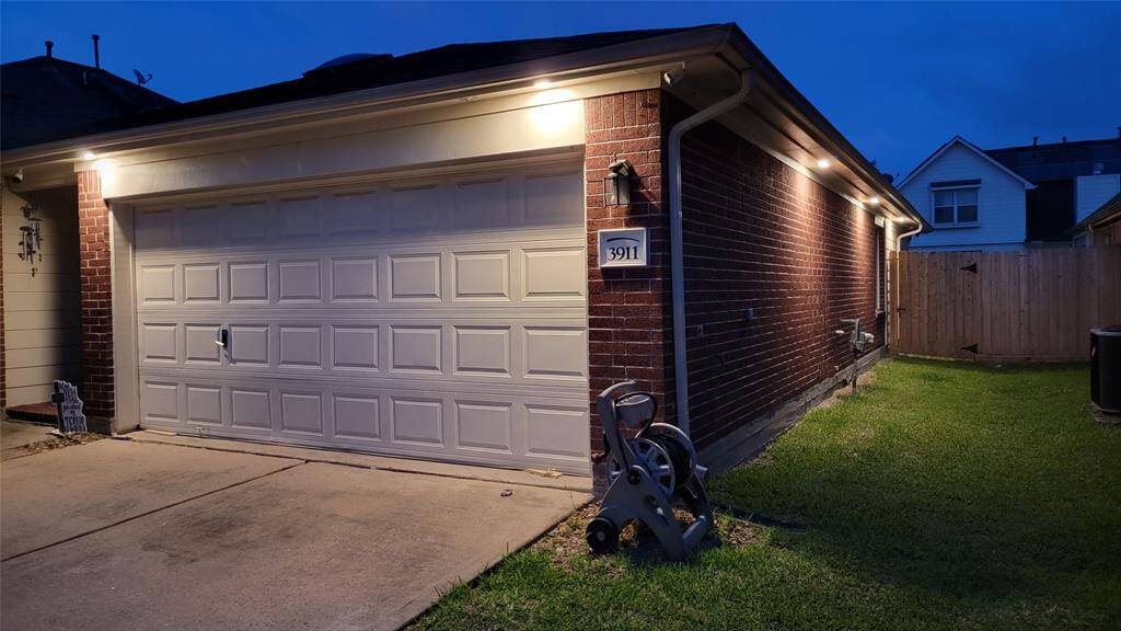 Photos taken at night to showcase the under eave lighting.  This photo shows a well-lit garage with a double door, brick exterior, and a neatly kept side yard. The driveway is concrete, and there's a wooden fence enclosing the backyard, creating a cozy and secure outdoor space.