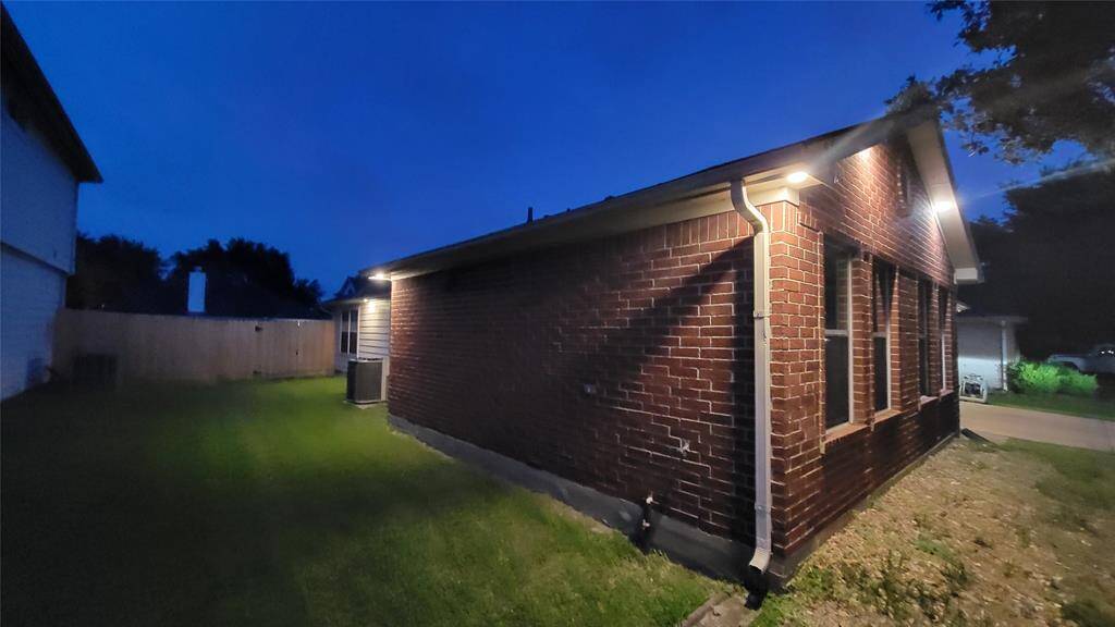 This photo shows the side of a brick house at dusk with exterior lighting. It features a small grassy yard and a wooden fence, creating a cozy and private outdoor space.