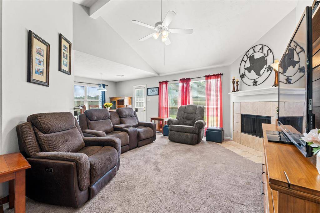This spacious living room features plush seatingj (furniture not included), a cozy fireplace, and large windows with red curtains offering plenty of natural light. The high ceiling with a fan and neutral decor create an inviting atmosphere, perfect for relaxation or entertaining.