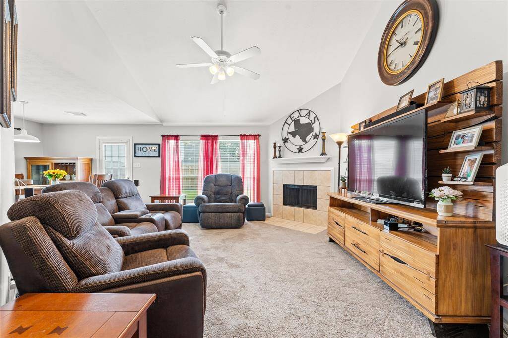 Spacious living room with high ceilings, cozy carpeting, and ample natural light. Features include a comfortable seating area, a large TV on a wooden entertainment unit, and a fireplace. Decorated with red curtains and a prominent wall clock, creating a warm, inviting atmosphere. (TV and decor not included)