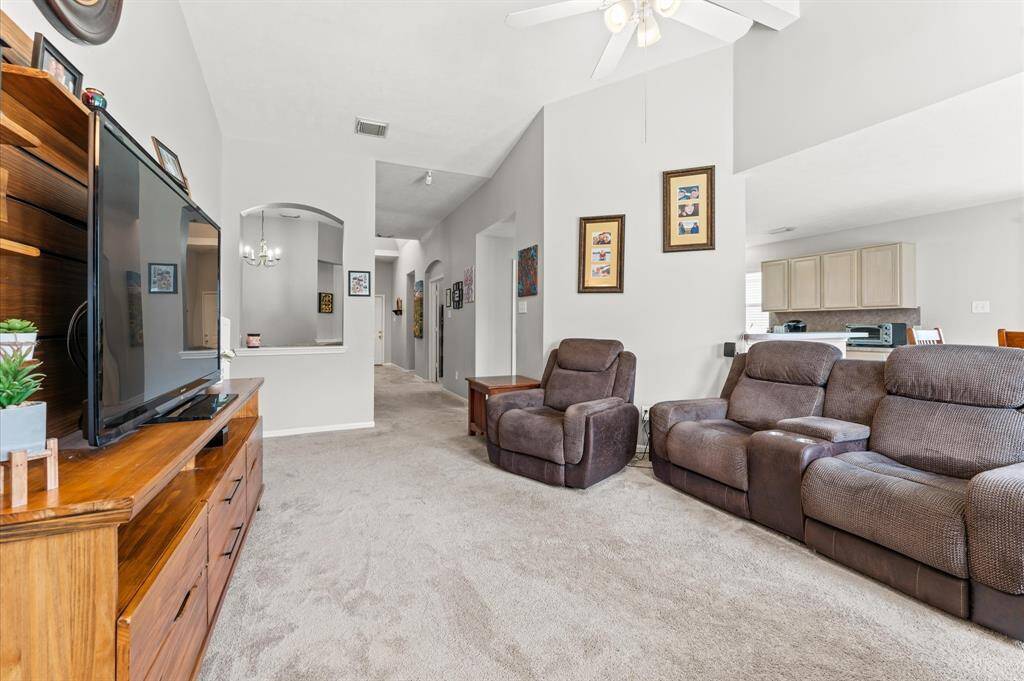 This inviting living room features cozy recliners, a large TV, and an open layout that flows into the kitchen. The neutral-toned walls and carpet create a warm, welcoming ambiance, complemented by decorative wall art and ceiling fans for comfort.  (decor, furniture, and TV not included)