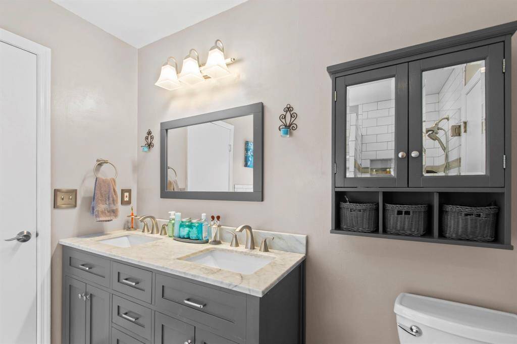 This bathroom features a modern double vanity with a marble countertop, under-mounted sinks, and elegant brushed nickel fixtures. It includes ample storage with a wall-mounted cabinet and decorative lighting, creating a stylish and functional space.
