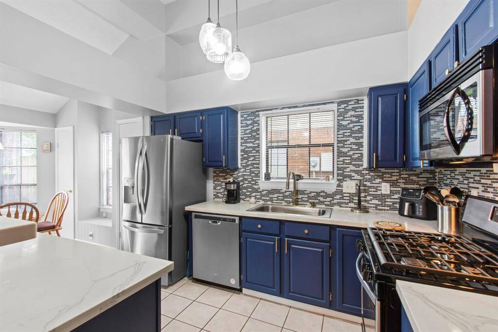 Modern kitchen with vibrant blue cabinets, stainless steel appliances, and a mosaic tile backsplash. Features a sleek island with Quartz countertops, pendant lighting, and ample natural light from multiple windows.