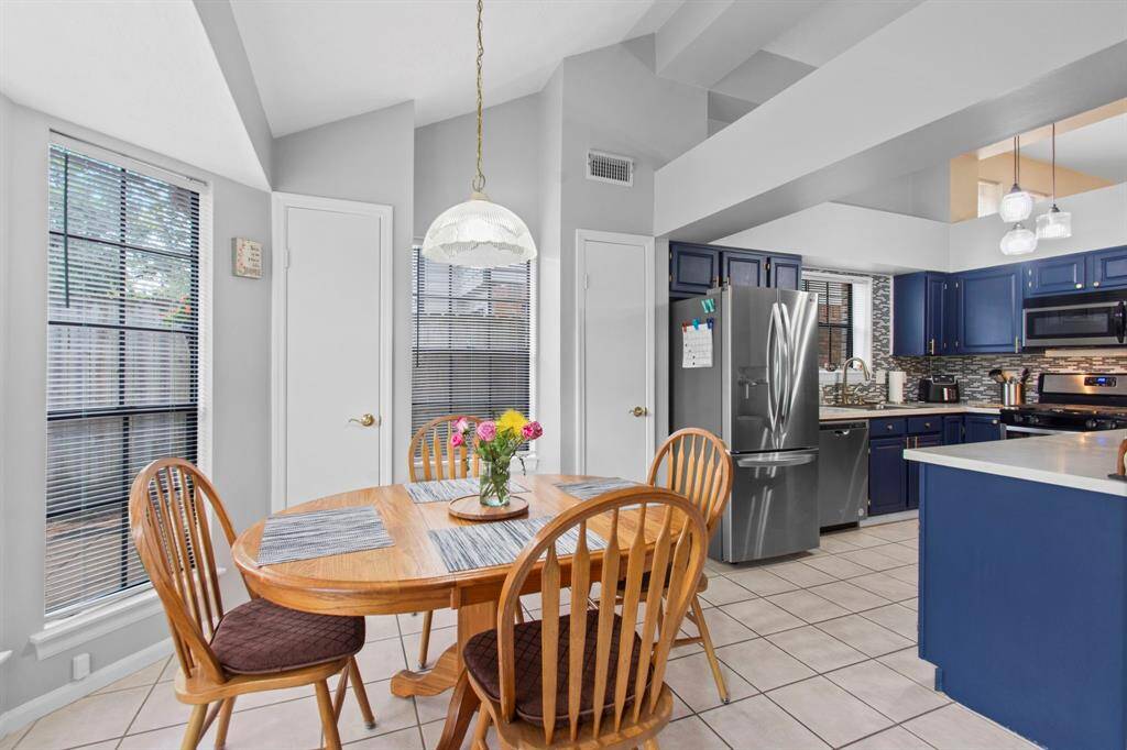 This kitchen features a modern, open layout with high ceilings and ample natural light. It includes stainless steel appliances, navy blue cabinetry, and a cozy dining area. The tile flooring adds a bright and clean look.