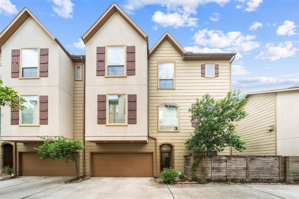 This is a modern, three-story townhouse with a beige and brown exterior, featuring large windows and a double garage. The property is surrounded by greenery, providing a welcoming and serene environment.