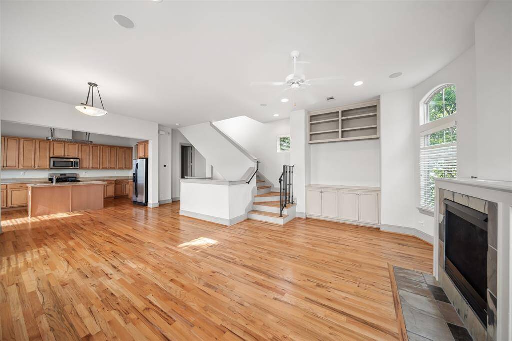 This spacious open-concept area features hardwood floors, a modern kitchen with ample cabinetry and stainless steel appliances, a cozy fireplace, and built-in shelves. The room is bright, with large windows and a staircase leading to the upper floor.