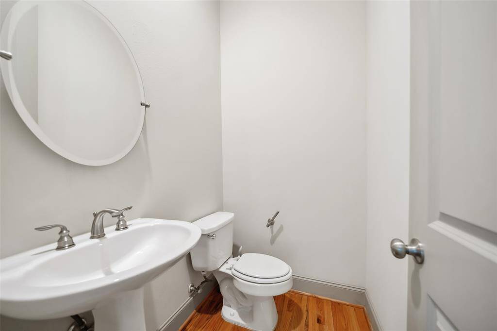 Compact half-bathroom with a pedestal sink, toilet, and round mirror. Features clean, white walls and a wood floor.