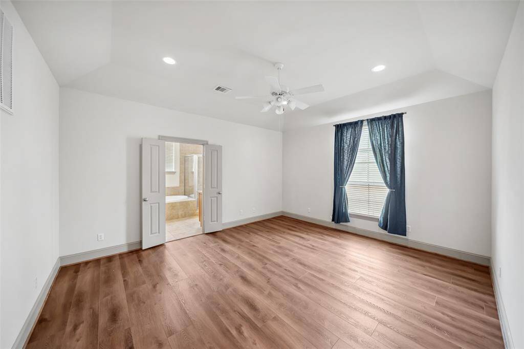 Spacious bedroom with wood flooring, recessed lighting, and ceiling fan. Features double doors leading to a bathroom and a window with blue drapes for natural light.