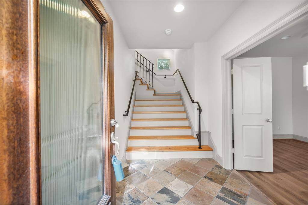 This entryway features a modern wooden door with glass panels, tiled flooring, and a wooden staircase. A small table with decorative items adds a welcoming touch.