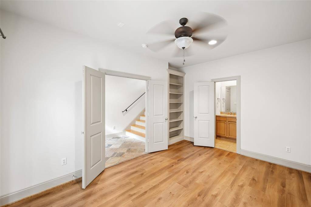 This bright room features hardwood floors, a ceiling fan, and built-in shelving. It has access to a staircase and an en-suite bathroom with wooden cabinetry, making it a versatile space for living or working.