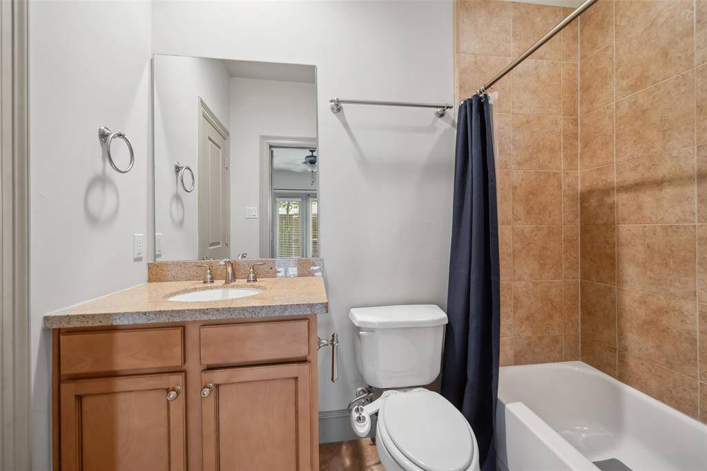 This bathroom features a sleek design with a single vanity, light wood cabinetry, and a granite countertop. It includes a toilet and a bathtub with a tiled shower area, accented by a dark curtain. Neutral tones create a clean, modern look.