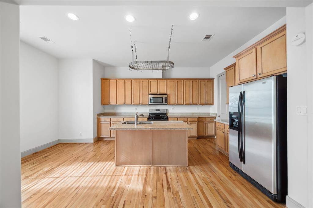 This kitchen features hardwood floors, light wood cabinetry, and a central island with a sink. It includes stainless steel appliances, ample counter space, and recessed lighting for a bright, modern feel.