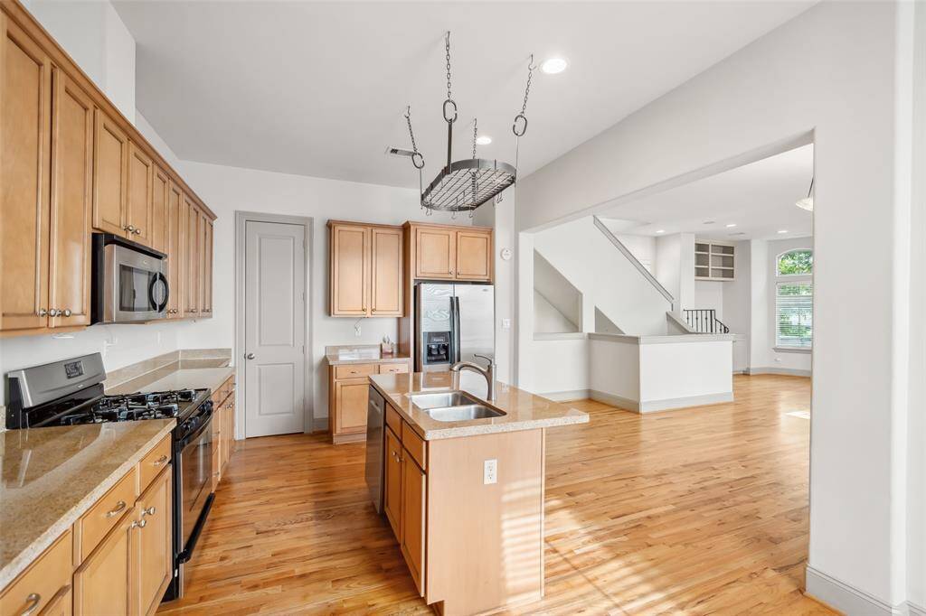 Bright open-concept kitchen with natural wood cabinets, stainless steel appliances, and granite countertops. The kitchen flows into a spacious living area with hardwood floors and large windows, offering ample natural light.