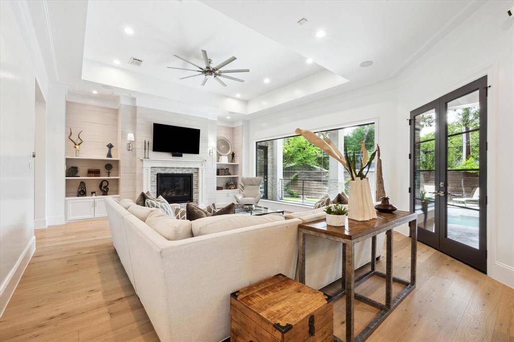This modern living room features a 42" fireplace with a shiplap mantle, custom built-in shelving, decorative wall sconces. It has abundant natural light from expansive custom plate windows and glass doors that open to a pool area, creating a seamless indoor-outdoor flow.