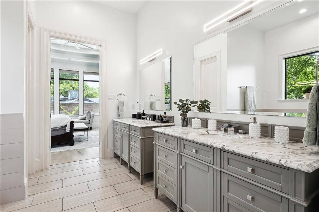 This spacious bathroom features dual vanities with marble countertops, modern lighting, and ample storage. The design flows seamlessly into a bright bedroom with large windows offering a view of the greenery outside.