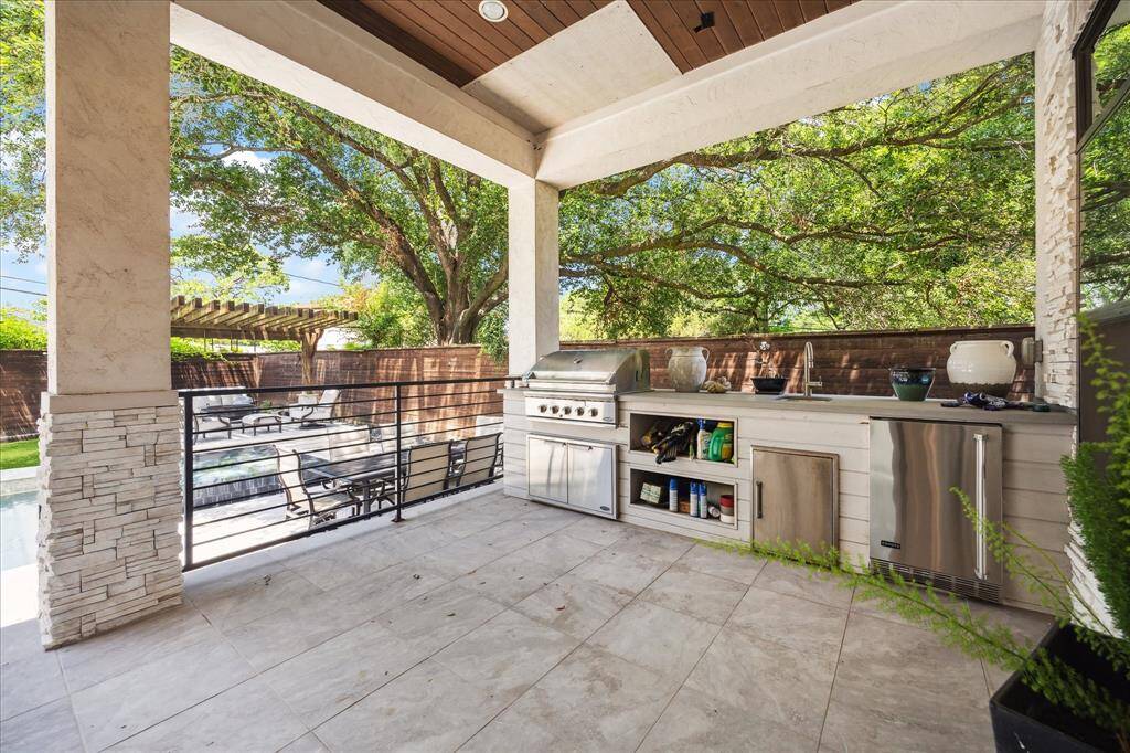 This photo showcases a stylish outdoor kitchen with a built-in grill, sink, and refrigerator, set on a covered patio with stone accents. It overlooks a landscaped backyard with a seating area and mature trees, sparkling pool, offering a perfect space for entertaining.