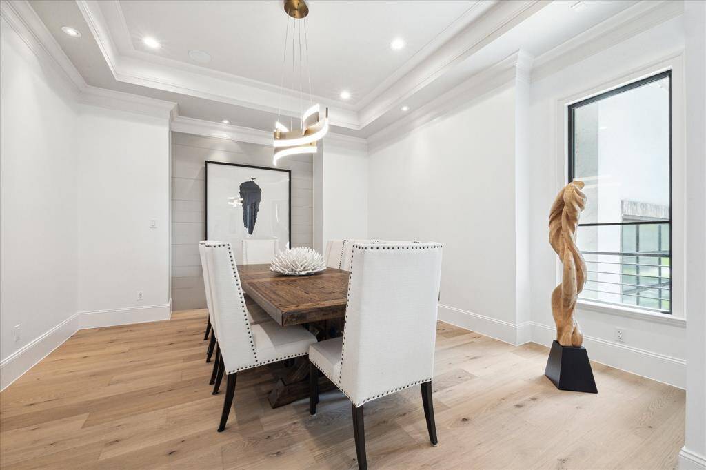 This elegant dining room features a modern chandelier, a tray ceiling with decorative moldings and a ship lap niche wall with recessed art lighting. The space is bright with natural light from a large window, complemented by French white oak wood floors.