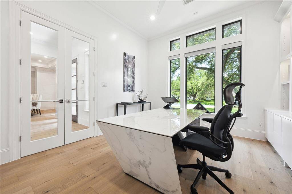 This bright, modern home office features built-in custom cabinetry with down lighting, large windows with a scenic view, hardwood floors, and elegant double glass doors, creating an inviting and productive workspace.