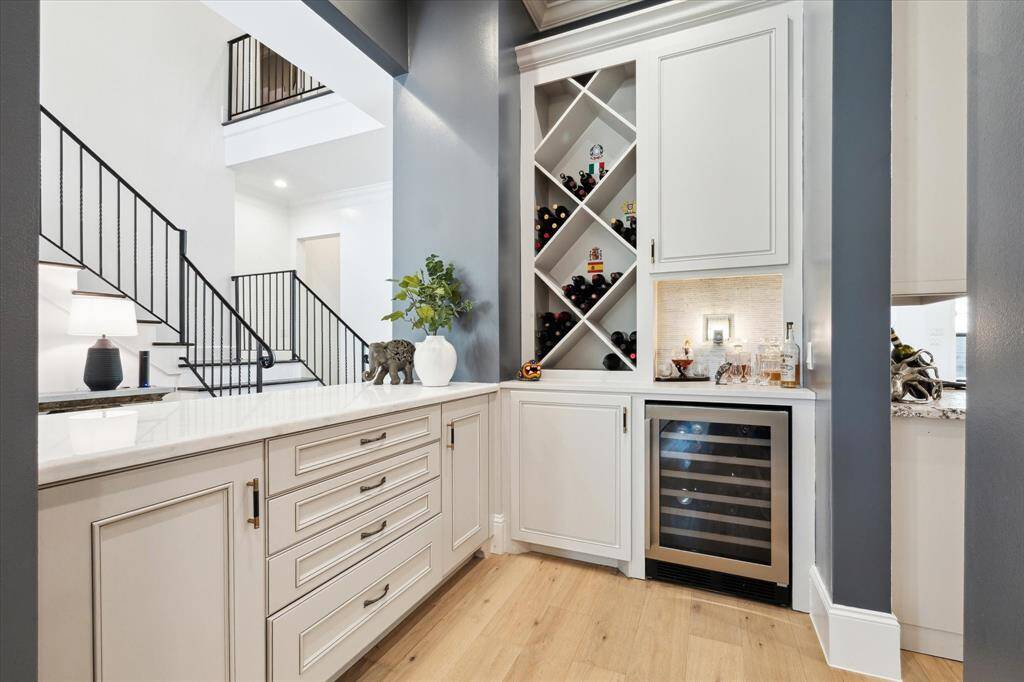 This photo showcases a stylish home bar area with built-in cabinetry, a wine rack, and a wine cooler. It features sleek design elements with a mix of white and dark tones, and an open layout that connects to an airy stairway. Perfect for entertaining and elegant storage.