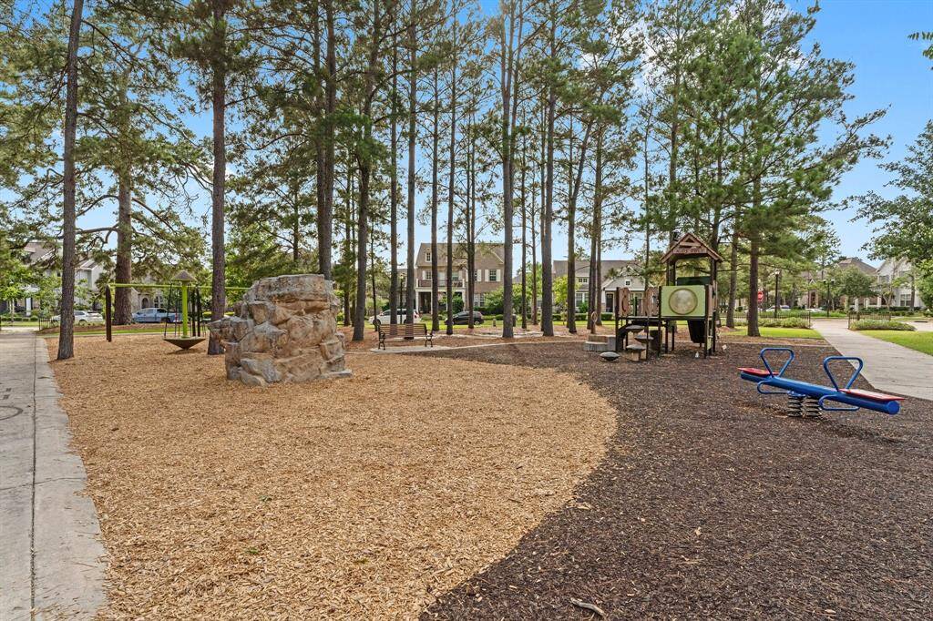 Spacious neighborhood playground surrounded by mature trees, featuring a climbing rock, play structures, and ample seating areas.