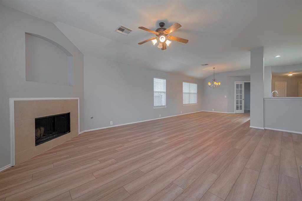 This spacious living area features wood-look flooring, a cozy fireplace, and neutral walls. It offers an open layout with ceiling fans and ample natural light from multiple windows. The room connects seamlessly to a dining area and kitchen, ideal for entertaining.
