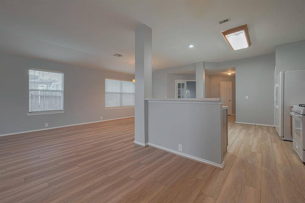 This photo showcases an open-concept living area with modern light wood flooring and neutral walls. The space features large windows for natural light, a partial dividing wall, and a visible kitchen area with appliances, creating a bright and airy atmosphere.