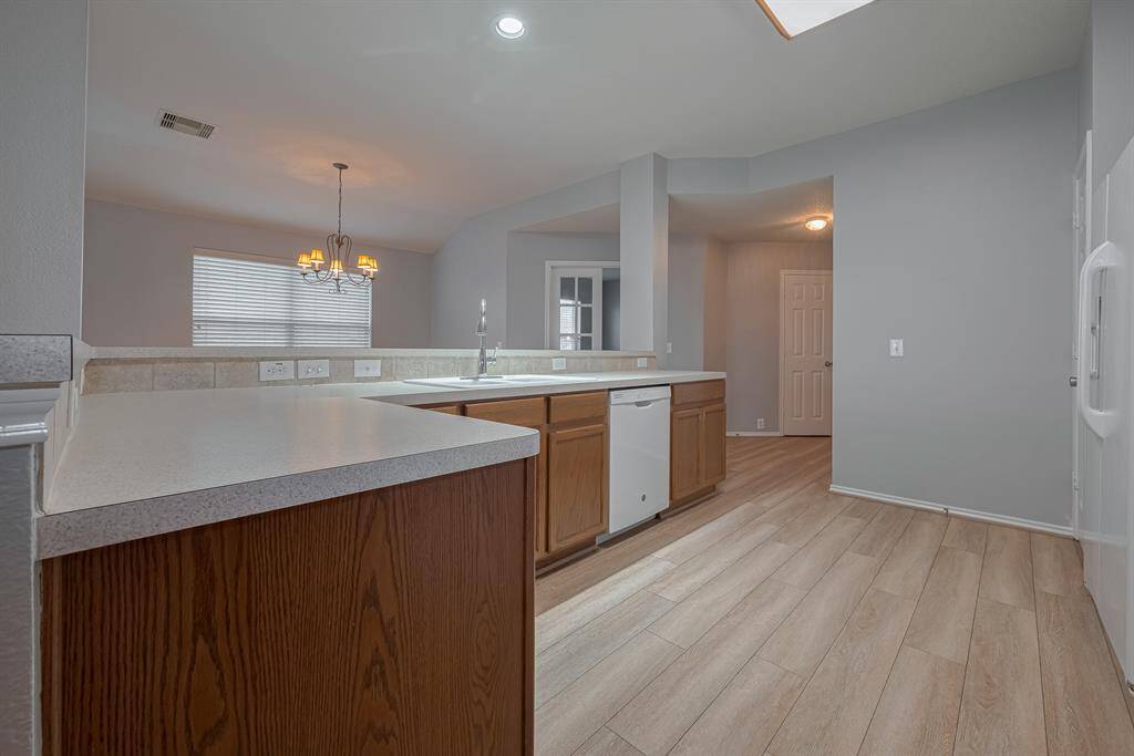 This kitchen features modern light wood flooring, a spacious countertop, and ample cabinetry. It includes a dishwasher and overlooks a dining area with a window and chandelier, creating a bright and inviting space.