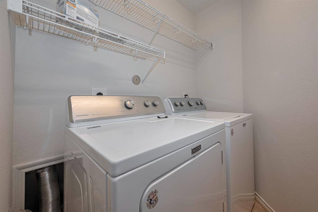 This photo shows a compact interior laundry room with a washer and dryer set. It features white walls, a light flooring, and a wire shelf above for storage.