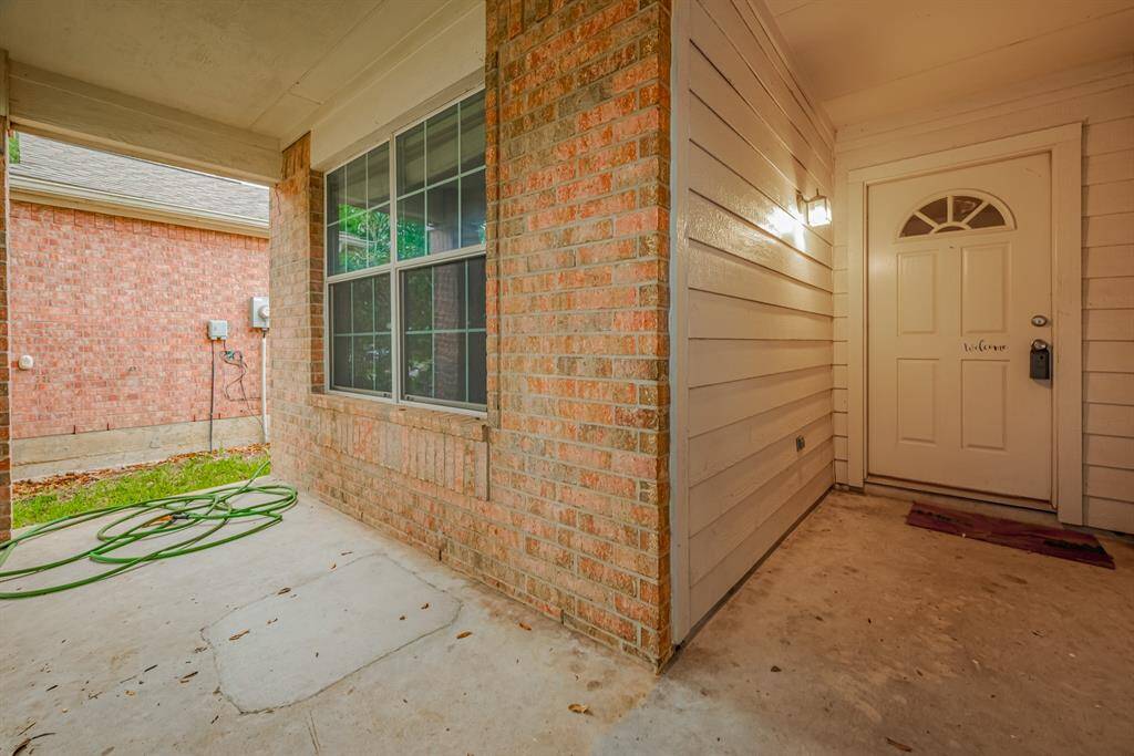 This photo shows a welcoming entryway with a covered porch. It features a brick wall, a large window, and a white front door with a small overhang. The area is well-lit and has space for outdoor decor.