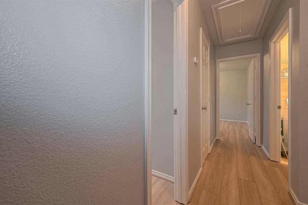 This photo shows a hallway with light wood flooring and neutral-colored walls. It features several doorways, leading to different rooms, and has a ceiling with a pull-down attic access. The area is well-lit, offering a clean and inviting appearance.
