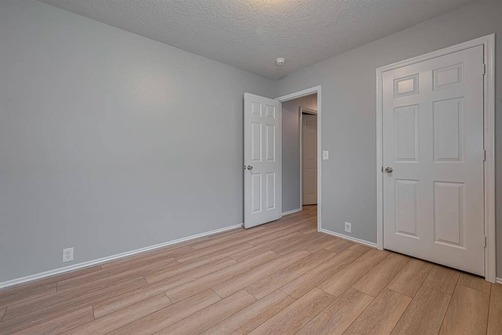 This is the primary bedroom with light gray walls and wood-look flooring. It features two white doors, one of which is open, leading to a hallway. The room has a simple ceiling light fixture.