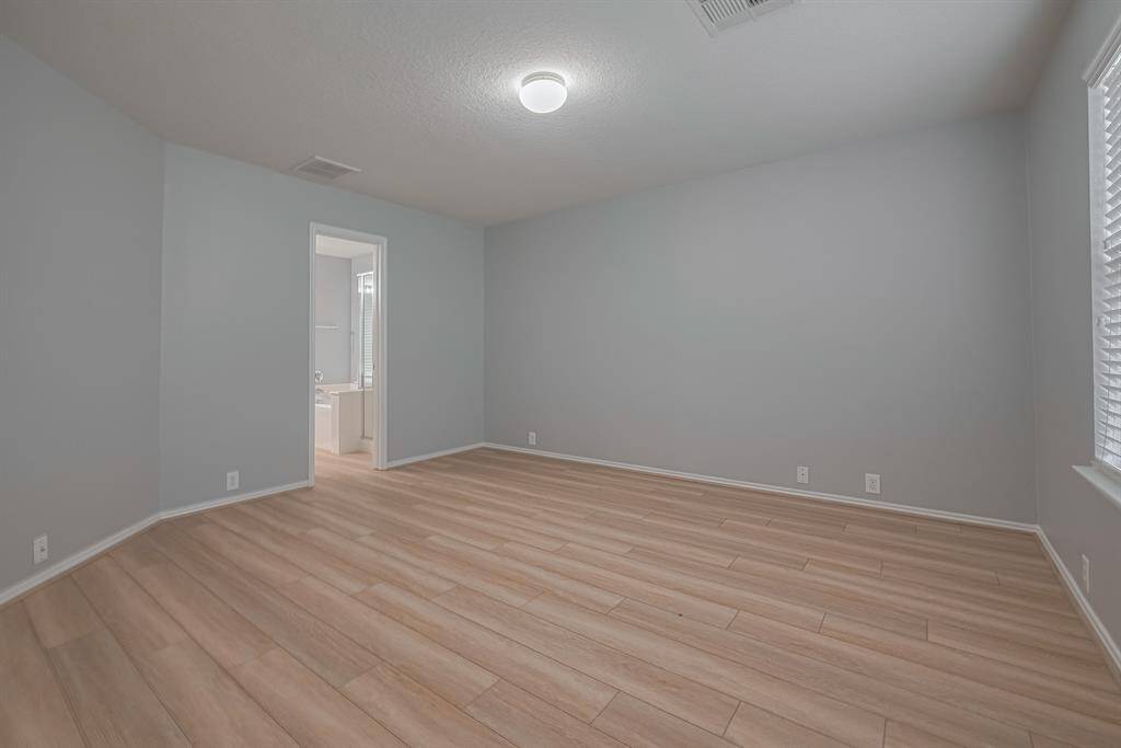 This photo showcases a spacious primary bedroom with light wood flooring and neutral walls, featuring a ceiling light and a window allowing natural light. There's an adjoining bathroom with a visible bathtub.