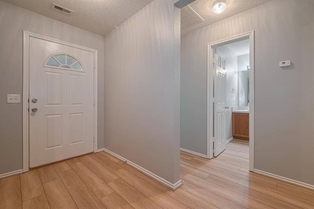 Bright entryway with light wood flooring, neutral gray walls, and a white front door. There's a hallway leading to a bathroom with visible vanity and mirror. The space feels clean and inviting.