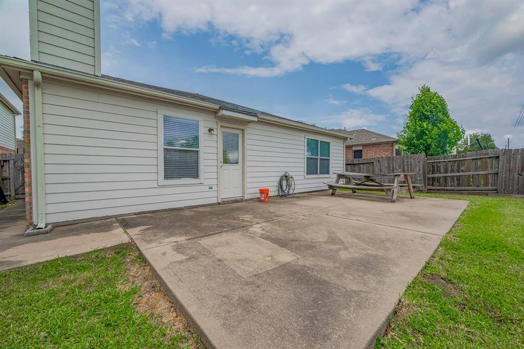This photo showcases a spacious backyard patio at the rear of a house, featuring a concrete surface ideal for outdoor activities. A wooden picnic table is present, and the area is enclosed by a wooden fence, offering privacy. The house exterior is light-colored with a door and windows leading to the patio.