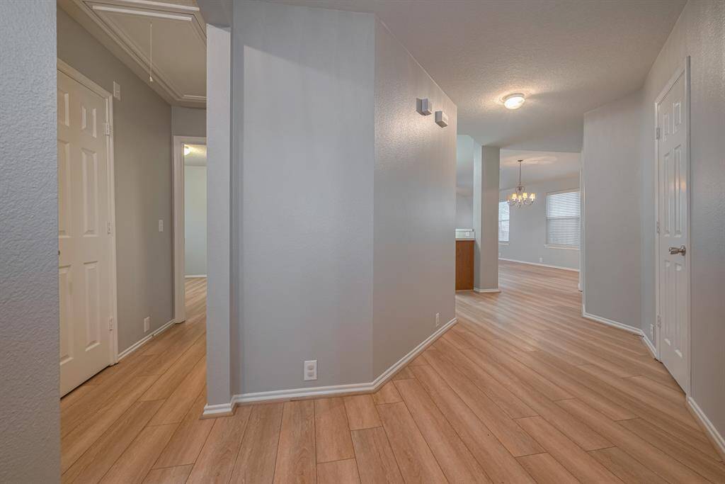 This photo features a bright hallway with light wood flooring and neutral gray walls. It leads into a spacious area with a chandelier, likely a dining room, providing an open and inviting atmosphere.