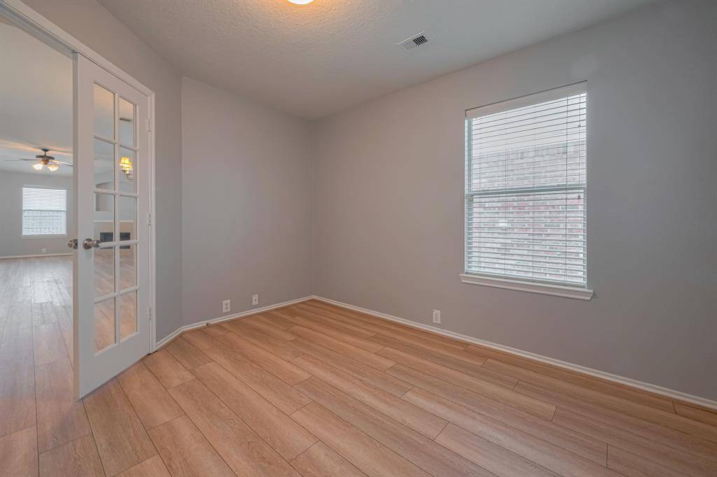 This photo showcases a bright, room with light wood flooring and neutral walls. It features a window with blinds, and a set of French doors leading to another room, allowing natural light to flow through. Ideal for an office or study.