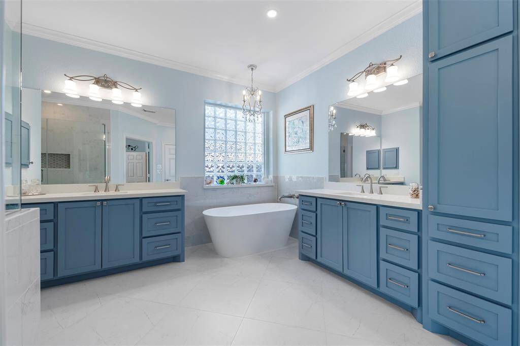 Luxurious primary bathroom boasting dual vanities, a sculptural soaking tub, and ample storage in rich blue cabinetry under soft pendant lighting.