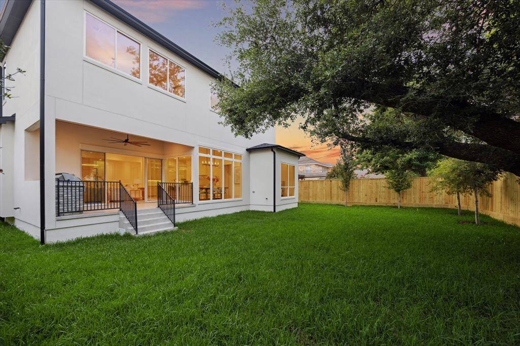 [BACKYARD] The backyard is a private oasis, framed by lush landscaping and showcasing the home’s stunning rear elevation. A wall of large windows offers a striking view of the interiors glowing warmly from within, creating a captivating blend of nature and architecture that enhances the home’s contemporary charm.