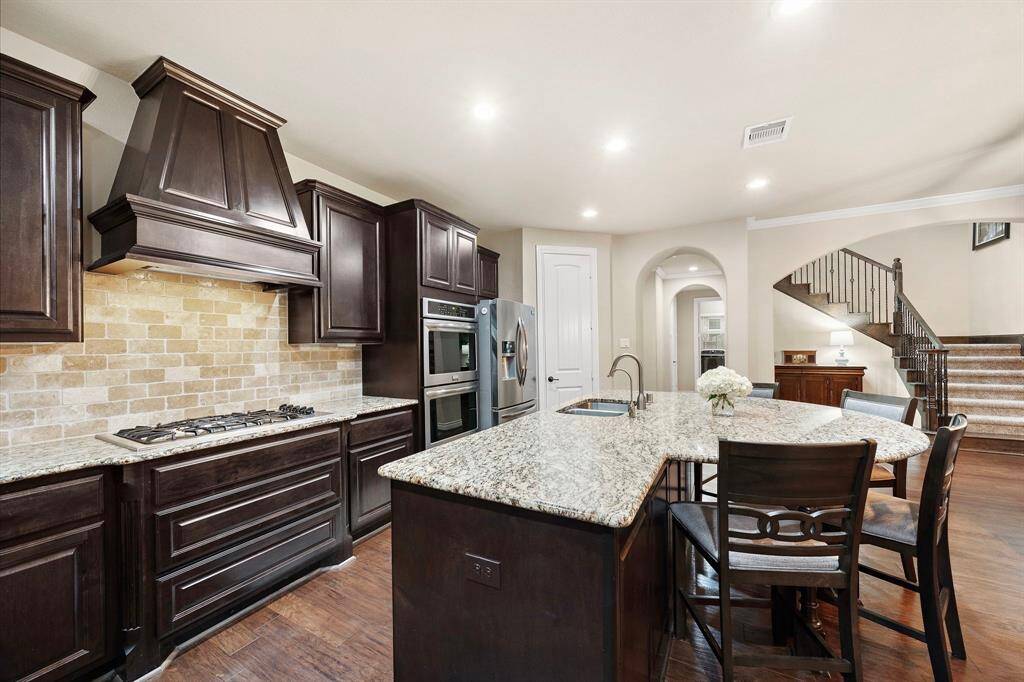 A five-burner gas cooktop, statement vent hood, and stone backsplash offer a refined culinary setup, while the oversized island adds storage and space to gather.