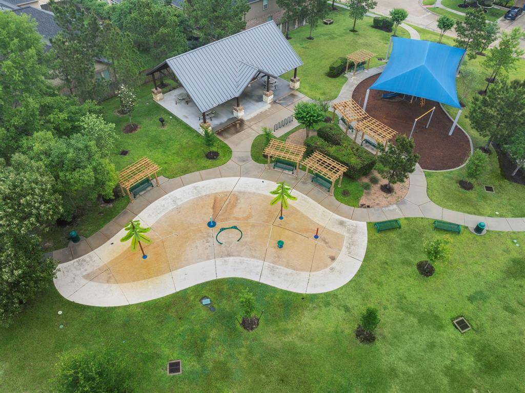 Community park includes a covered pavilion with picnic tables, a shaded playground with mulch surfacing, and a splash pad featuring interactive water elements. Multiple benches and trellised seating areas offer convenient spots to relax and enjoy the lush green space.