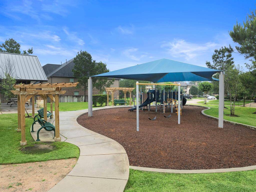 Shaded playground features a covered play structure with swings and slides, surrounded by mulch for comfort and safety. Nearby pergola seating and paved walkways offer convenient spots to relax, making this neighborhood park a welcoming space for outdoor enjoyment.